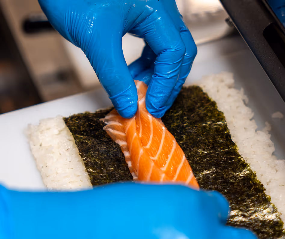 Hands with blue gloves placing salmon on rice and seaweed.
